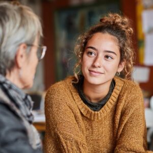 University student wearing a warm brown sweater smiling while conversing with a peer in a relaxed environment.