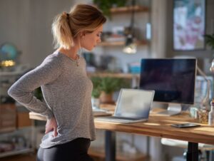 Woman holding her lower back in pain while working at a standing desk in a home office setting.
