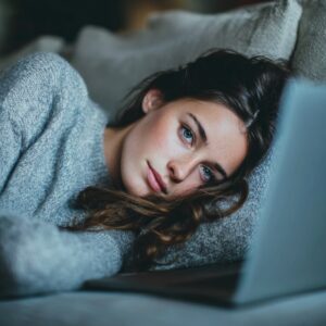 Young woman lying on a sofa in a grey sweater, using a laptop and gazing thoughtfully.