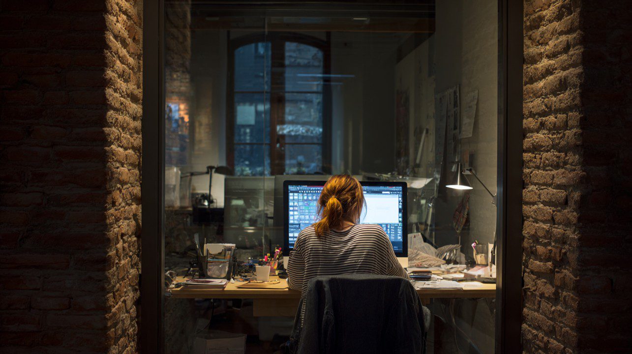 Woman working late at her desk in a small brick-walled office in Barcelona, lit by a desk lamp.