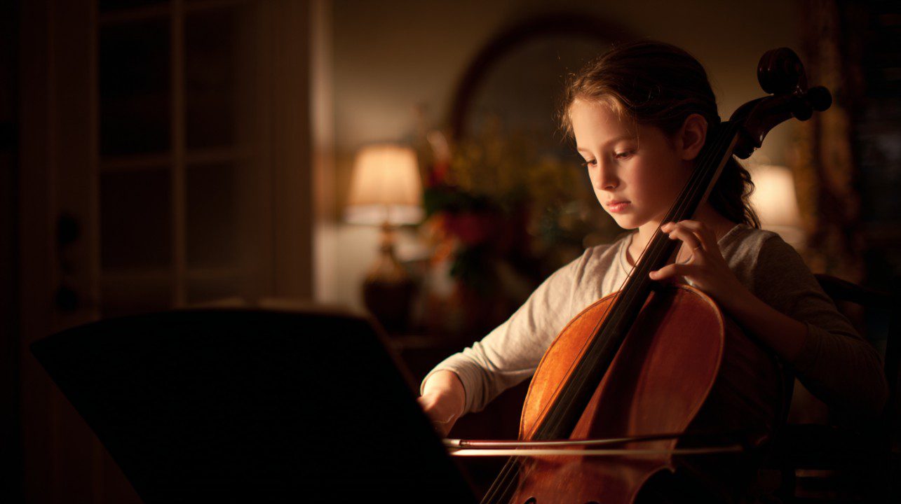 Young girl plays the cello while reading sheet music in a warmly lit room.