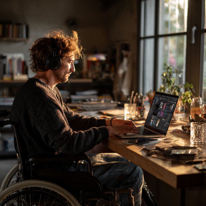 Young man in a wheelchair wearing headphones working on a laptop in a cozy, sunlit home office.