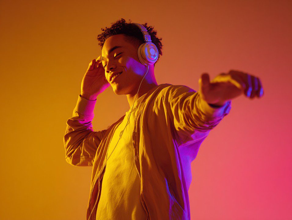 Young man with headphones enjoying music under neon orange and pink lighting, wearing a casual jacket and smiling.