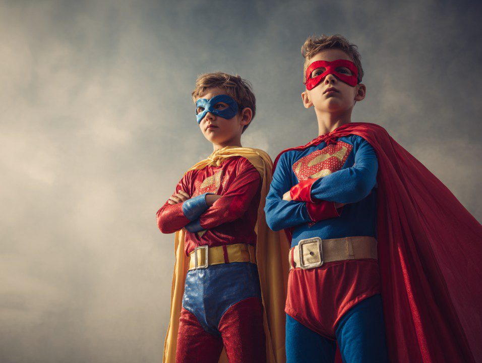 Two boys dressed in superhero costumes with masks and capes, standing confidently against a cloudy sky.
