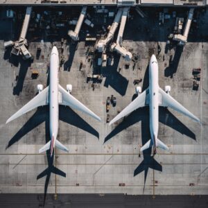 Aerial photograph of two large commercial airplanes parked at an airport terminal with jet bridges and service vehicles in view.