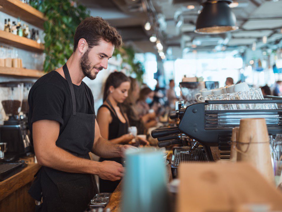 Smiling barista prepares coffee behind a busy café counter, with customers and coworkers in a vibrant coffee shop setting.