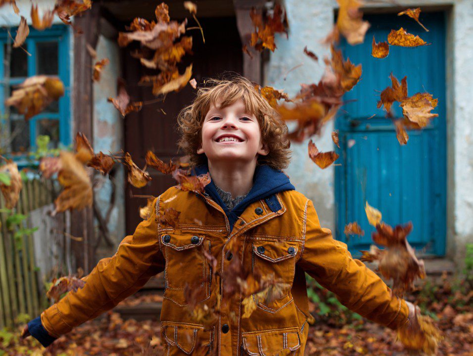 Cheerful boy in an orange jacket smiling and throwing autumn leaves in the air in a rustic outdoor setting.