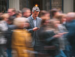A businessman checks his phone while a crowd rushes past, creating a blur effect that symbolizes focus and digital distraction in a fast-paced urban setting.