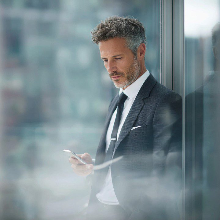 Mature businessman in a suit standing by a glass window and checking his phone, representing modern communication in a corporate setting.