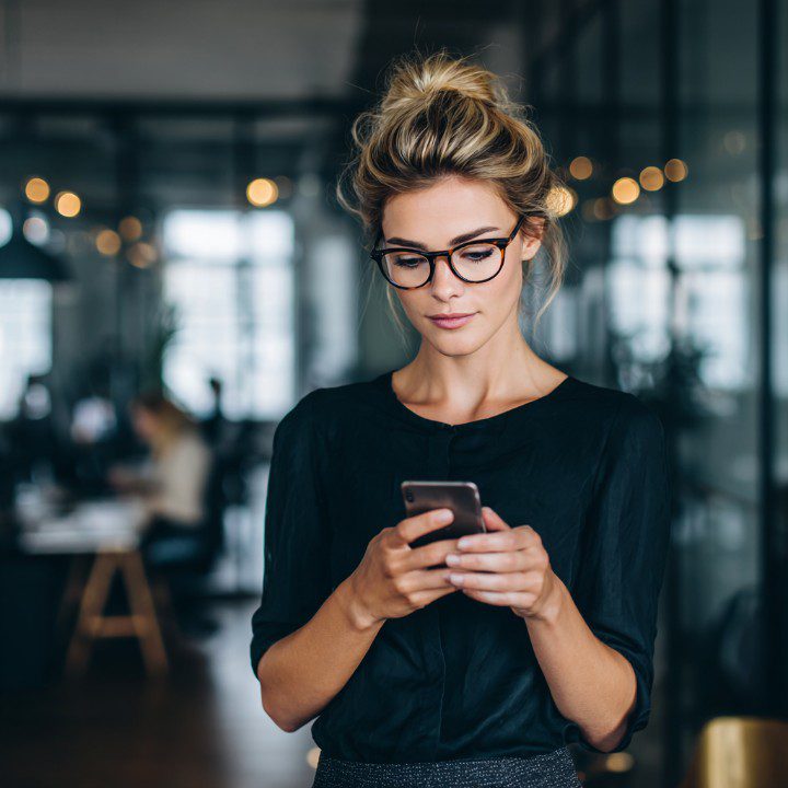 Young professional woman in glasses using smartphone in modern open-plan office, symbolizing communication and productivity.