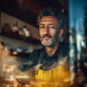 Confident male café owner in yellow apron behind a window, holding a smartphone and smiling warmly.