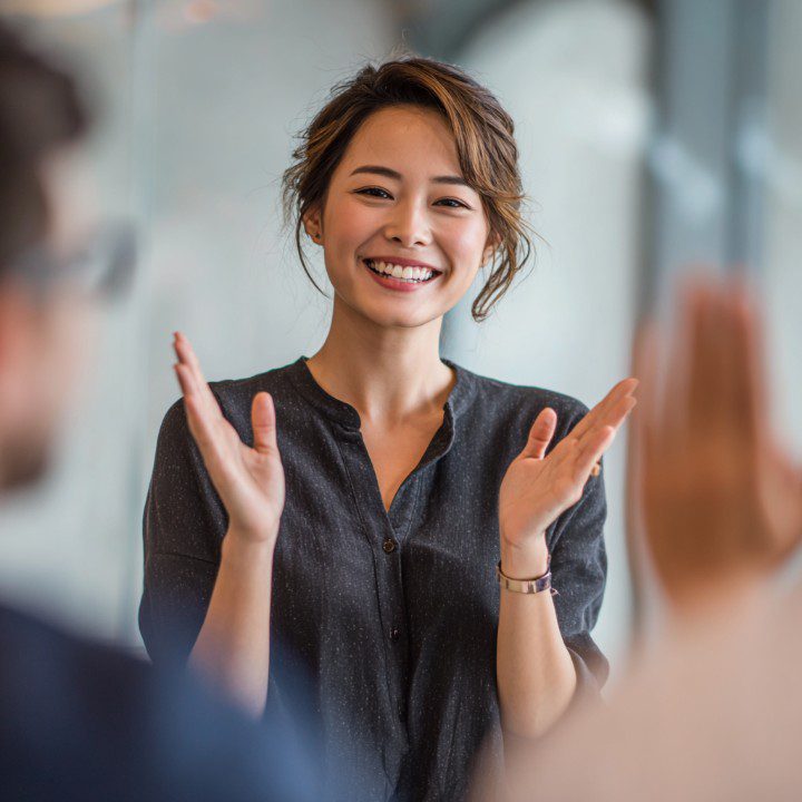 Cheerful Asian businesswoman applauding and smiling while speaking to a colleague during a team meeting in a modern office.