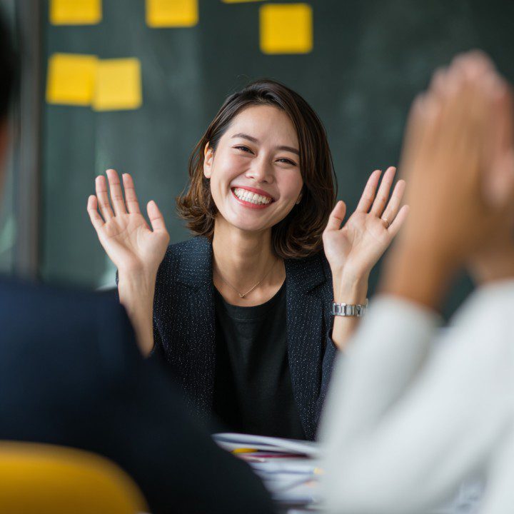 Cheerful Asian businesswoman smiling and waving during a friendly team meeting in a modern office setting.