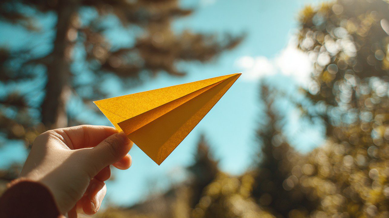 A child’s hand holding a yellow paper airplane against a bright blue sky with trees in the background.