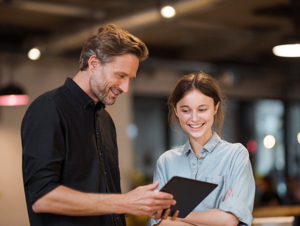 Two smiling colleagues collaborate using a tablet in a modern office setting, showcasing teamwork and professional communication.