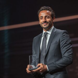 A smiling man in a suit holding a clear award trophy on stage, representing corporate recognition and professional success.