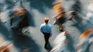 A man in a light blue shirt walking alone through a fast-moving, blurred crowd in an urban setting, symbolizing solitude and focus.