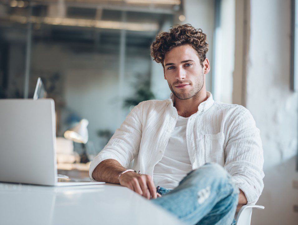 Young man in a white shirt and jeans sitting confidently at a desk with a laptop in a modern office environment.
