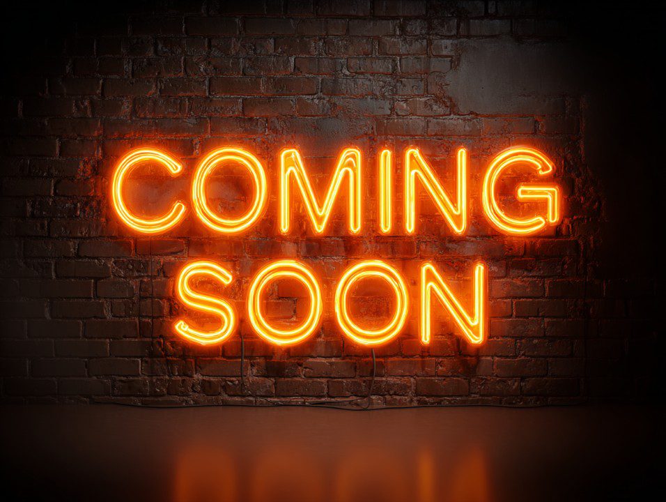 Realistic glowing orange neon sign reading "COMING SOON" on a dark brick wall background.
