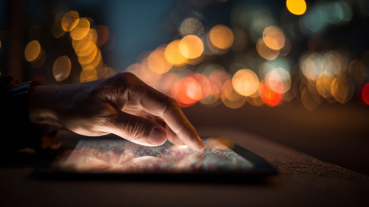 A close-up of a hand scrolling on a digital tablet at night, with a glowing bokeh background of city lights.