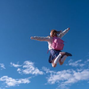 Carefree schoolgirl with pink backpack jumping high with arms outstretched against a bright blue sky, representing joy and youth.