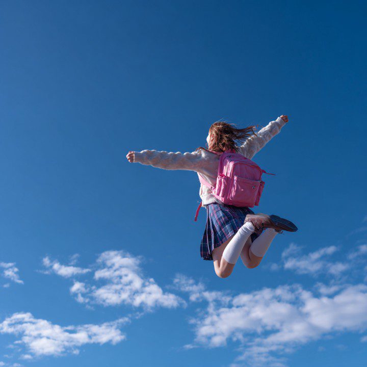 Carefree schoolgirl with pink backpack jumping high with arms outstretched against a bright blue sky, representing joy and youth.