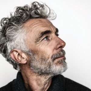 Close-up profile of an older man with curly gray hair and a beard, expressing wisdom and maturity against a white background.