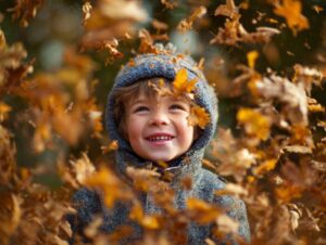 Smiling young boy in a grey wool coat and hat, surrounded by falling golden autumn leaves in an outdoor setting.