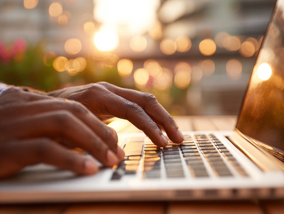 Close-up of dark-skinned hands typing on a laptop keyboard with warm bokeh lights in the background.