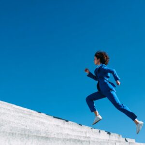 A young woman in a stylish blue suit running up concrete stairs under a clear blue sky, symbolizing motivation, success, and leadership.