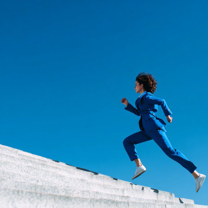 A young woman in a stylish blue suit running up concrete stairs under a clear blue sky, symbolizing motivation, success, and leadership.