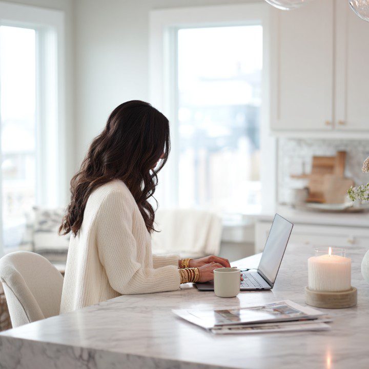 Woman with long dark hair working on a laptop in a bright, cozy, minimalist kitchen with a candle and coffee.
