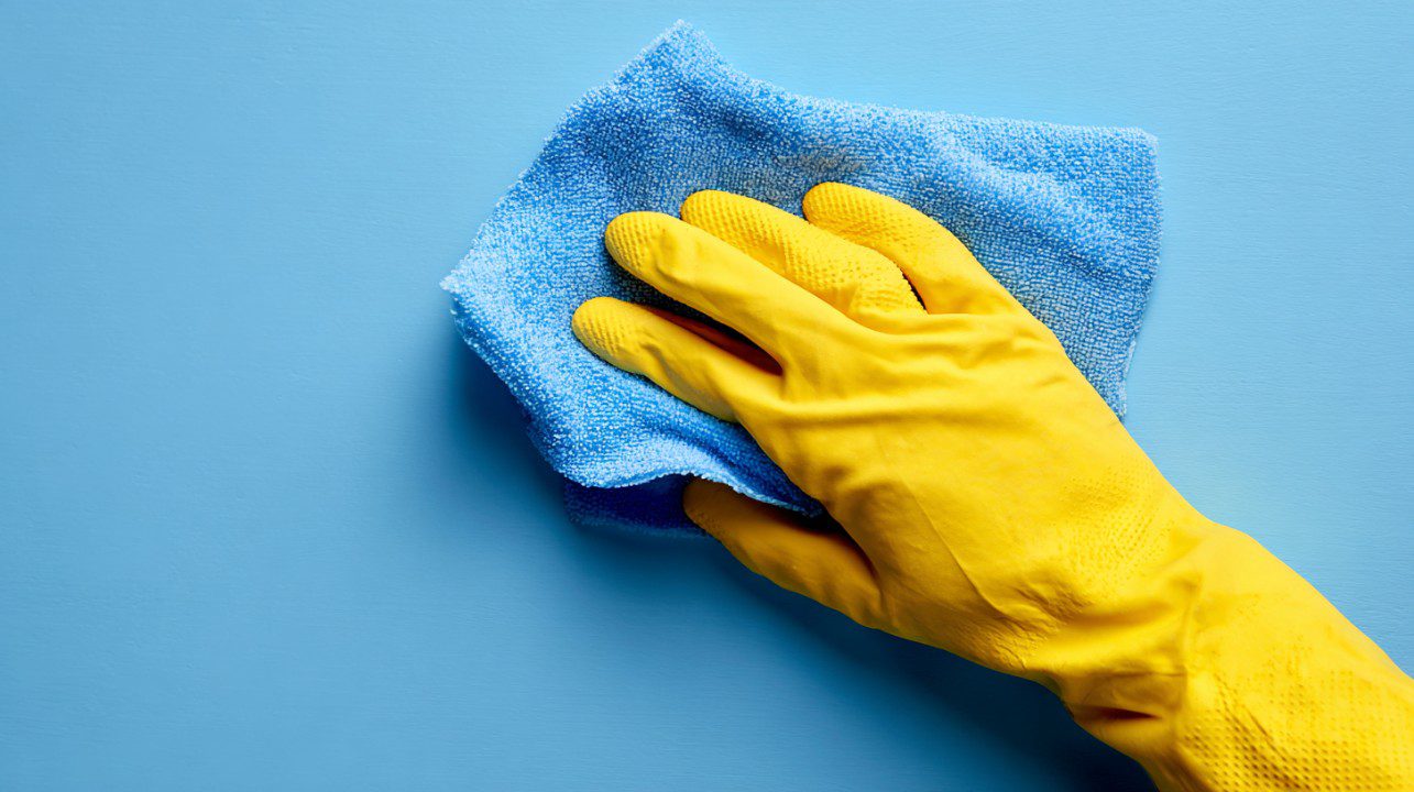 Close-up of a hand wearing a yellow rubber glove cleaning a blue surface with a microfiber cloth, symbolizing hygiene and sanitation.