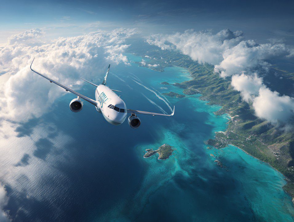 Airplane flying above turquoise tropical ocean and lush island coastline under cloudy skies, viewed from a high aerial angle.