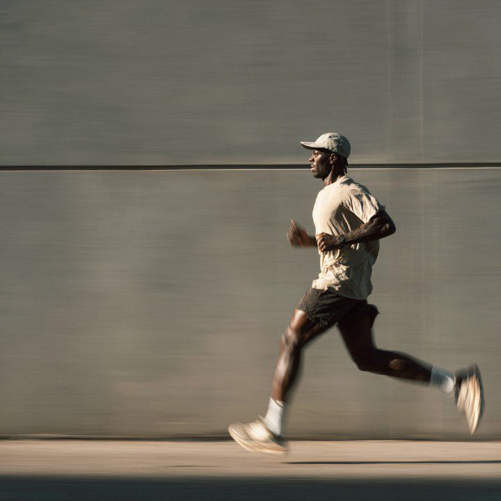 Full-body side view of athletic man running outdoors along a minimalist gray wall, symbolizing motion, fitness, and determination.