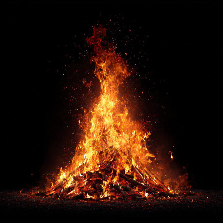 Tall bonfire with vivid orange flames and sparks rising against a solid black background, creating a dramatic and energetic scene.