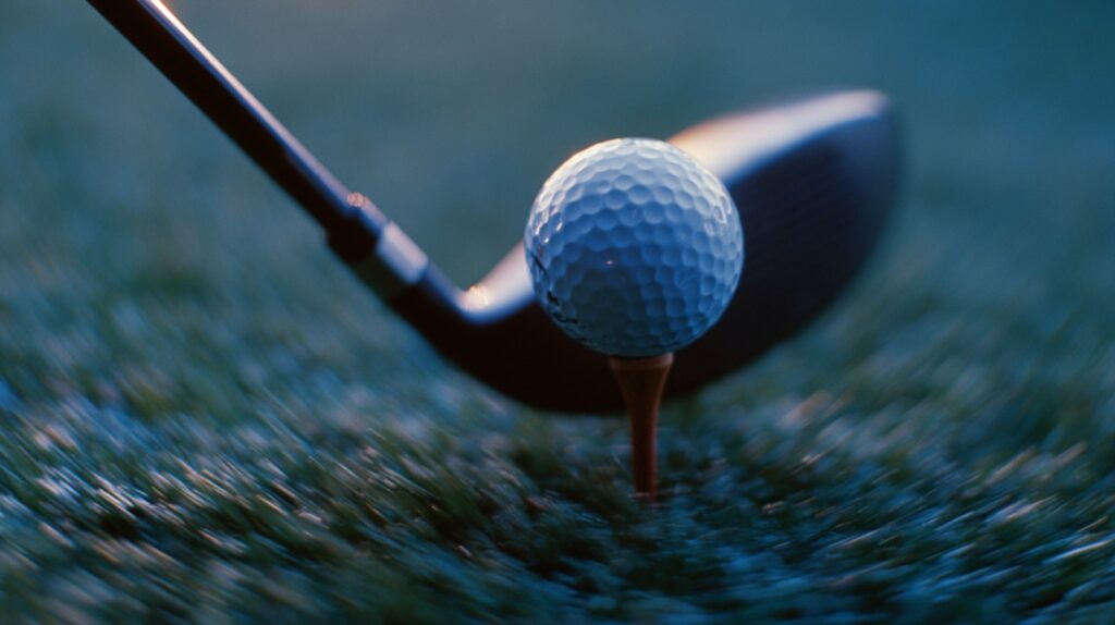 Close-up photo of a golf ball on a tee with a driver about to swing, set against a lush green field.