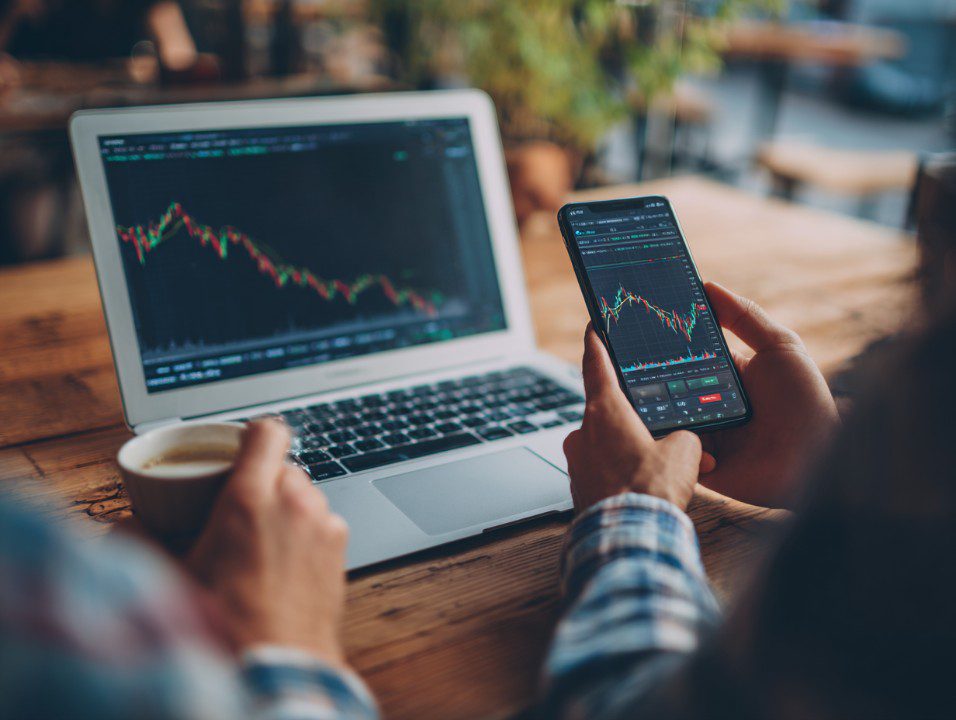 A couple using a smartphone and laptop to review live stock market charts while seated at a wooden table, with coffee in hand.