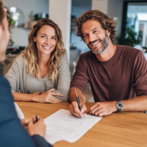A smiling couple sits at a desk signing a real estate contract with a professional agent, representing homeownership and financial planning.