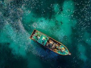 Drone image of a fishing trawler surrounded by a large school of fish in bright turquoise ocean water.