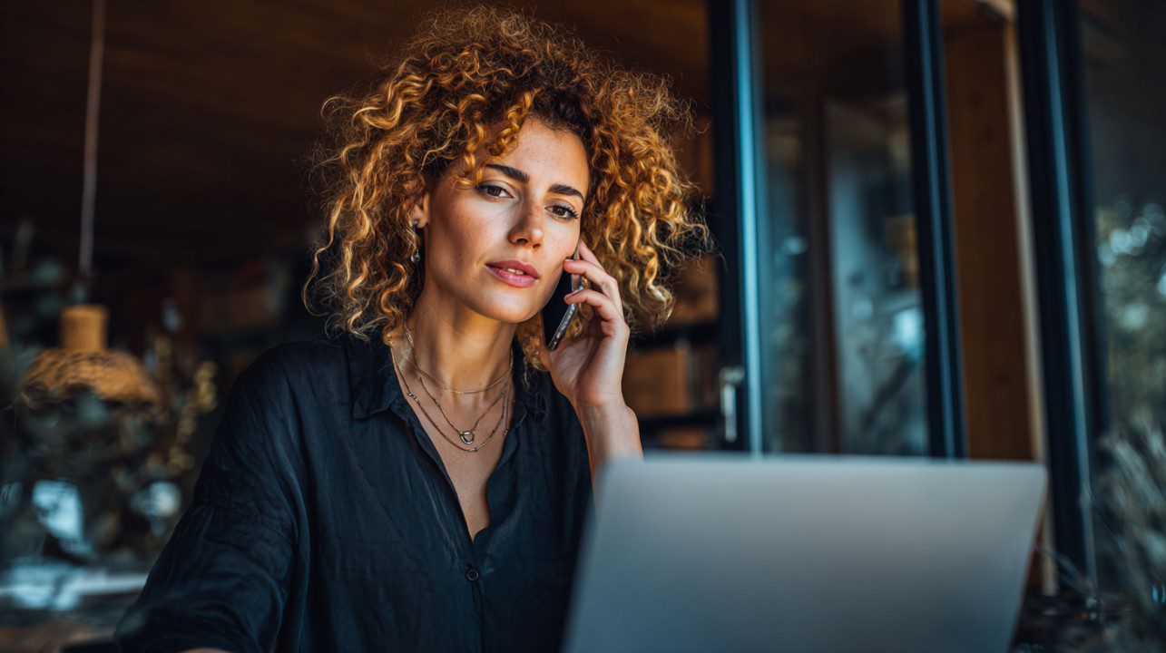 Focused entrepreneur talking on the phone while working on a laptop in a modern home office, representing business productivity and remote work.