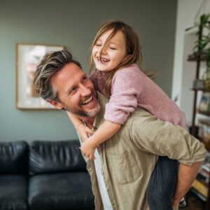 Happy father giving his daughter a piggyback ride in the living room, both laughing and enjoying quality family time at home.