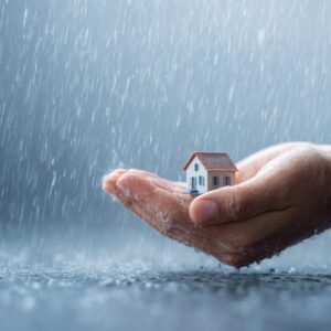 Close-up of a hand cradling a small house model under heavy rain, symbolizing protection, shelter, and home insurance.