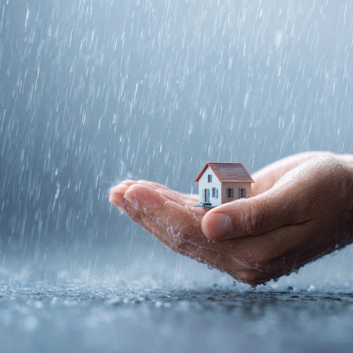 Close-up of a hand cradling a small house model under heavy rain, symbolizing protection, shelter, and home insurance.