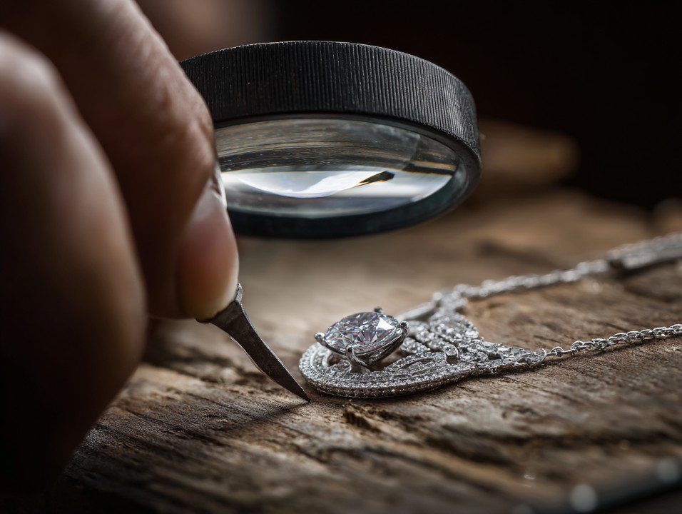 A jeweler uses a magnifying glass and fine tool to inspect a detailed diamond necklace on a wooden workbench, representing craftsmanship and luxury.