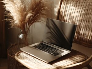 Sleek laptop on a wicker chair in a cozy interior with warm lighting and pampas grass, ideal for creative workspace mockups.