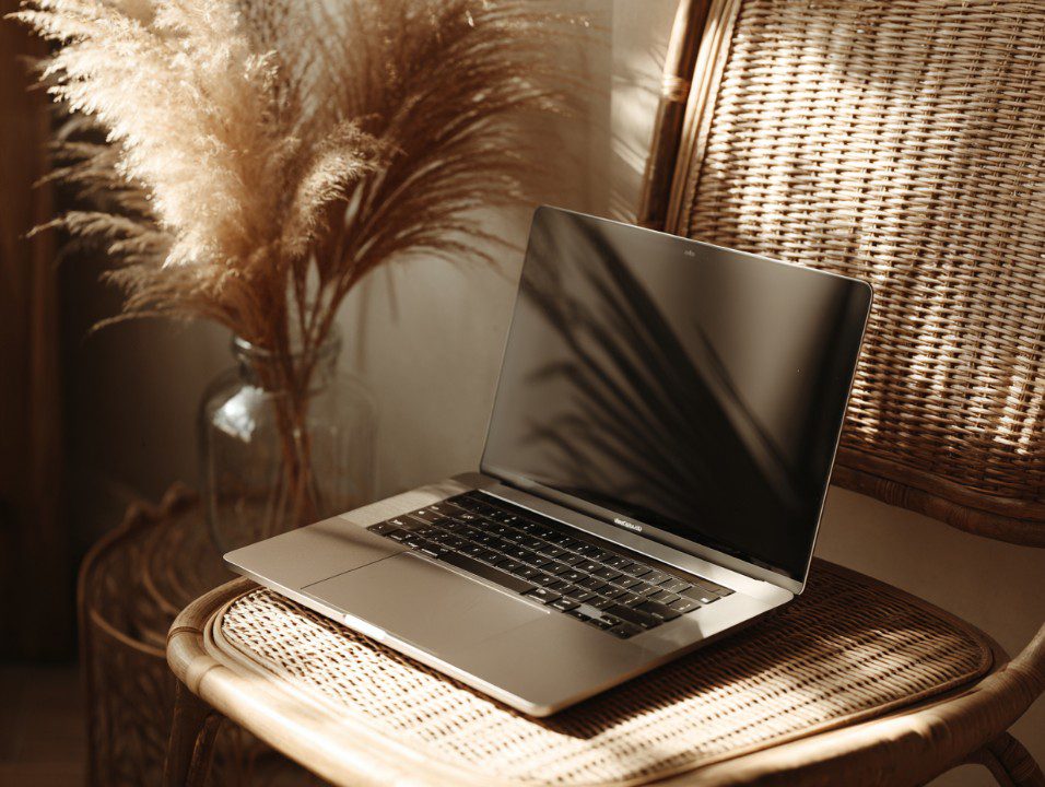 Sleek laptop on a wicker chair in a cozy interior with warm lighting and pampas grass, ideal for creative workspace mockups.