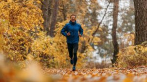 Middle-aged man in blue jacket jogging along a forest trail covered in golden autumn leaves, expressing health and seasonal activity.