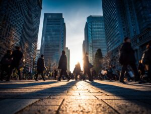 Silhouettes of morning commuters walking through Marunouchi business district in Tokyo, Japan, with tall glass skyscrapers and sun flare in the background.