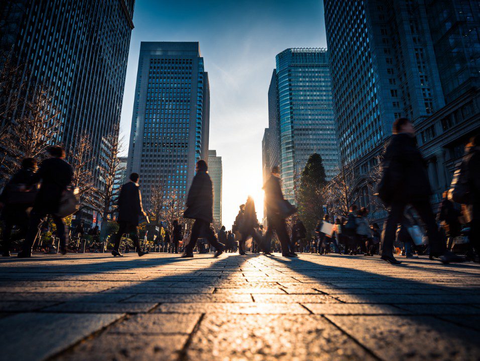 Silhouettes of morning commuters walking through Marunouchi business district in Tokyo, Japan, with tall glass skyscrapers and sun flare in the background.
